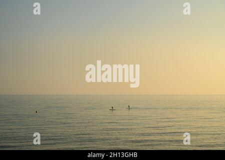 Zwei Paddlebarder, die vor Sonnenuntergang in sanftem Oktoberlicht mit Gelb- und Blautönen durch ruhiges Wasser fahren. East Sussex, England. Stockfoto