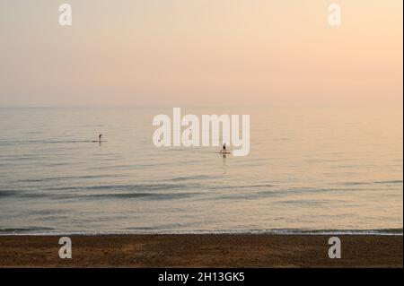 Zwei Paddlebarder, die im Oktober am Hove Beach in East Sussex, England, in sanften Rosa- und Blautönen durch ruhiges Wasser fahren. Stockfoto