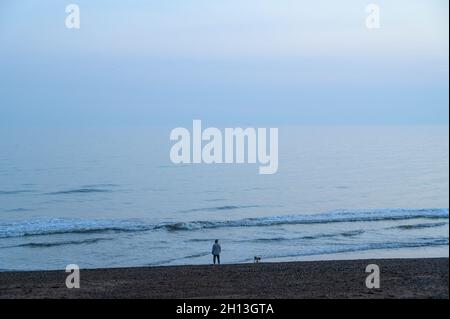 Blaue Stimmung: Eine junge Frau und ihr Hund am Ufer des Hove-Strandes blicken auf das ruhige Meer bei Sonnenuntergang. East Sussex, England. Stockfoto