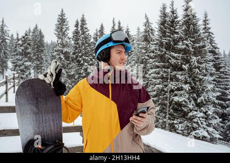 Wintersport und Erholung, Freizeitaktivitäten im Freien. Mann in Skiausrüstung, trägt eine Schutzbrille mit Smartphone . Snowboarder Mann beim Wandern Stockfoto