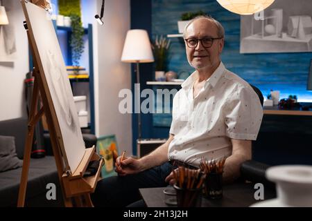 Porträt eines älteren kaukasischen Künstlers, der in einem Kunststudio sitzt. Älterer Mann mit künstlerischer Phantasie, der während der Arbeit an der Vasenzeichnung die Kamera anschaut. Gealterte Person mit Meisterwerk auf Leinwand Stockfoto