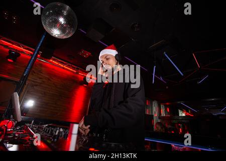 Frohes Neues Jahr und frohe Weihnachten Charismatischer Discjockey in rotem weihnachtsmann Hut, Kopfhörer und Hoody spielt Musik an DJ-Plattenspielern. Party im neuen Jahr Stockfoto