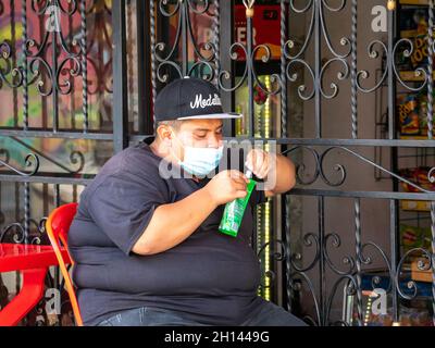 Medellin, Antioquia, Kolumbien - Dezember 23 2020: Ein fetthaliger Mann mit einer Maske sitzt auf einem roten Plastikstuhl und öffnet einen Beutel mit einigen Lebensmittelsachen Stockfoto