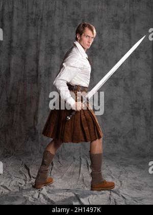 Ein junger Mann in einem Lederkilt und einer weißen Schnürbluse. Ein schottischer Ritter mit einem zweihändigen Schwert. Foto in voller Länge im Studio auf einem grauen Hintergro Stockfoto