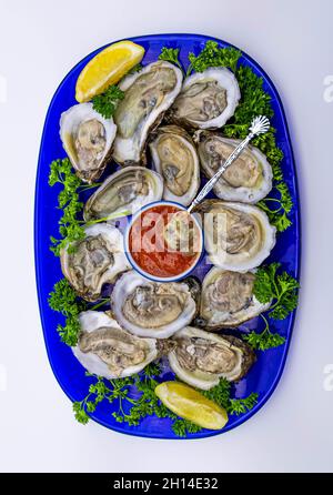 Frische, rohe Austern, die auf der Halbschale auf einem blauen Glasplatte mit roter Cocktailsauce und farbenfrohen Garnierungen serviert werden. Stockfoto