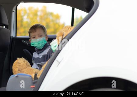 Ein siebenjähriger netter Junge mit medizinischer Maske sitzt an einem warmen, sonnigen Herbsttag während der Coronavirus-Pandemie in einem Auto. In der Nähe befindet sich ein Spielzeugbär. Auswahl Stockfoto
