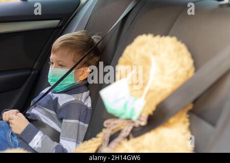 Ein siebenjähriger netter Junge mit medizinischer Maske sitzt an einem warmen, sonnigen Herbsttag während der Coronavirus-Pandemie in einem Auto. In der Nähe befindet sich ein Spielzeugbär. Auswahl Stockfoto