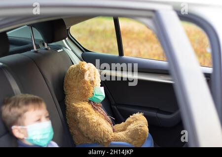 Ein siebenjähriger netter Junge mit medizinischer Maske sitzt an einem warmen, sonnigen Herbsttag während der Coronavirus-Pandemie in einem Auto. In der Nähe befindet sich ein Spielzeugbär. Auswahl Stockfoto