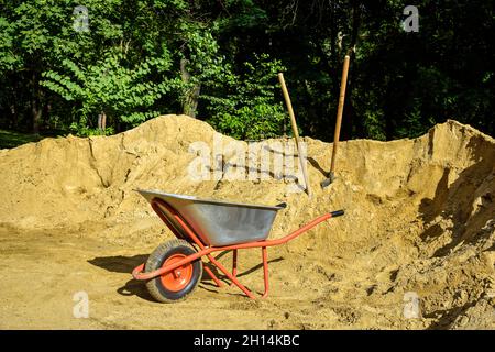 Bau-Schubkarre auf dem Hintergrund von Sandhaufen. Schaufeln im Sand stecken. Bauarbeiten im Park. Nahaufnahme. Selektiver Fokus. Stockfoto