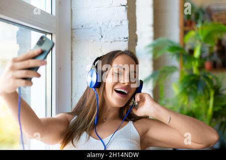 Eine junge Frau mit Kopfhörern singt morgens zu Hause. Portrait einer attraktiven jungen Frau in Kopfhörern mit Telefon - Apps zum Musikhören Stockfoto