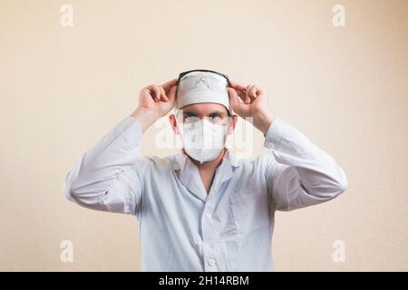 Arzt in weißem Labormantel und schützender Atemmaske passt seine Schutzbrille isoliert auf den gelben Hintergrund an. Stockfoto