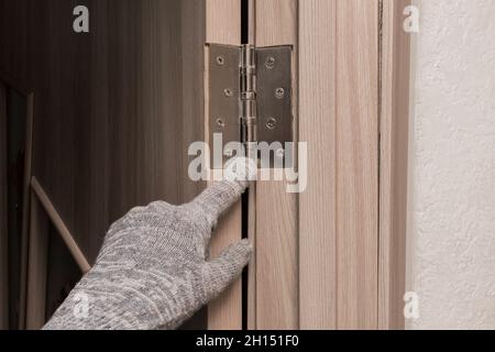Die Hand eines Arbeiters in einem Bauhandschuh zeigt mit dem Finger auf eine Holztür, die sich mit einem eisernen Türscharnier öffnet. Stockfoto