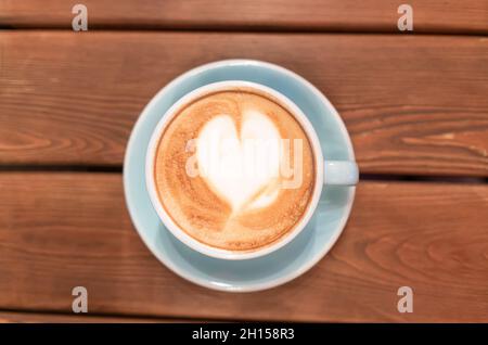 Herz aus Kaffeeschaum in einer blauen Tasse auf einem Holztisch, Draufsicht. Cappuccino oder Mokka am Morgen in einem Café. Latte Art Stockfoto
