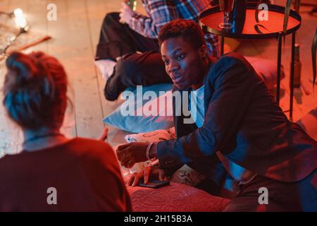 Afrikanischer junger Mann spricht mit einer jungen Frau, während sie auf den Kissen auf dem Boden auf einer Party liegen Stockfoto