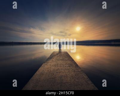 Mann, der allein auf dem Pier stand und den Sonnenuntergang sah, spiegelte sich im ruhigen Seewasser wider. Malerische Aussicht mit einer einsamen Person auf dem Dock Blick auf den Horizont Stockfoto