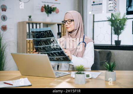 Kompetente Praktizierende in Hijab, die die Ergebnisse des Röntgenscans untersuchte, während sie mit einem kabellosen Laptop am Tisch saß. Arabischer Arzt auf der Suche nach einer Art der Patientenbehandlung. Stockfoto