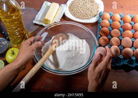 Hände schneiden Zitrone mit Messer auf Holzbrett. Hausmannskost-Konzept. Behälter mit Mehl, Eiern, Butter und Kochzutaten auf Holztisch. Stockfoto