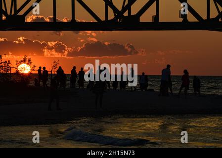 Touristen treffen sich unter der Mackinac Bridge, einer der längsten Brücken der Welt über die Straße von Mackinac, bei Sonnenuntergang, Mackinaw City, Michigan, USA Stockfoto