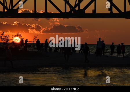 Touristen treffen sich unter der Mackinac Bridge, einer der längsten Brücken der Welt über die Straße von Mackinac, bei Sonnenuntergang, Mackinaw City, Michigan, USA Stockfoto