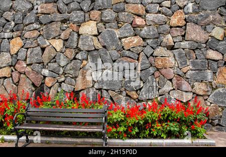 Foto einer abgenutzten Bank aus Holz und Stahl mit roten Blumen und Steinwänden im Hintergrund. Stockfoto