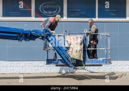 Weißrussland, Bobruisk - 02. September 2020: Zwei Bauleute, die auf einer Hebebühne in der Nähe der Fassade eines Geschäfts in der Stadt arbeiten, entfernen alte Werbespender Stockfoto