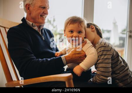 Älterer Mann mit seinen zwei Enkeln. Älterer Mann, der mit zwei Jungen zu Hause auf einem Stuhl sitzt. Stockfoto