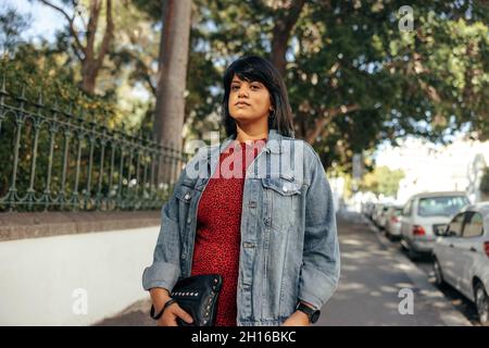 Frau mittleren Erwachsenen, die auf einem Bürgersteig in der Stadt steht. Selbstbewusste Frau, die neben einem Park steht und die Kamera anschaut. Frau in Denim ja Stockfoto