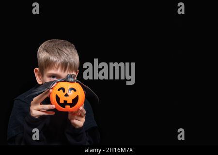 Ein Kind, das einen orangefarbenen Kürbis-förmigen Korb mit einem grinsenden Gesicht, Jacks Laterne und einer Fledermaus auf schwarzem Hintergrund hält. Der Junge wartet auf Halloween Stockfoto