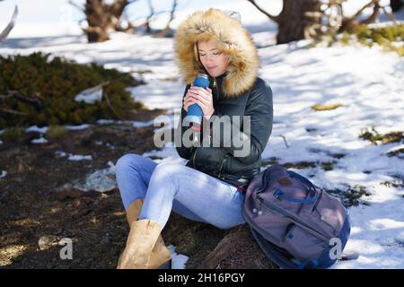 Frau, die etwas heißes aus einer Thermosflasche aus Metall trinkt, die auf einem Felsen in den verschneiten Bergen sitzt. Stockfoto