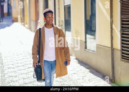 Ein junger schwarzer Mann, der die Straße entlang läuft, trägt eine Aktentasche und ein Smartphone. Stockfoto