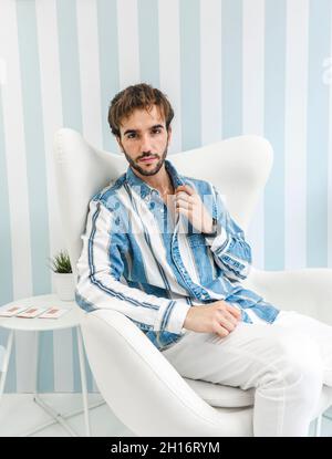 Eleganter bärtiger Kerl, der die Kamera und den verstellenden Kragen des T-Shirts anschaut, während er auf einem weißen Sessel sitzt Stockfoto