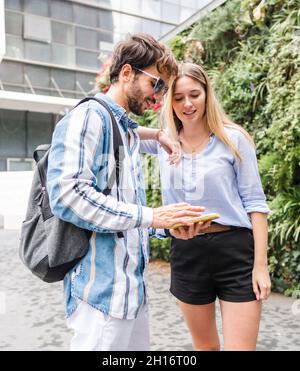 Frau in legerer Kleidung und Mann mit Rucksack stehend und Blick auf den Bildschirm des Smartphones mit einem Lächeln im Universitätshof Stockfoto