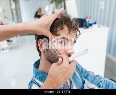 Von oben anonyme weibliche Meister schneiden männlichen Client, während er auf die Kamera im Friseurladen schaut Stockfoto