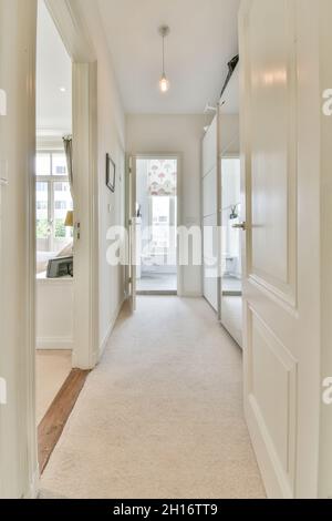 Enger leerer Korridor mit verspiegeltem Kleiderschrank, der an sonnigen Tagen zum Wohnzimmer und Bad in einem modernen, geräumigen Apartment führt Stockfoto