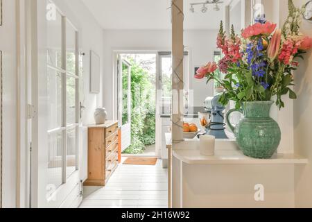 Blühende Blumen in grüner Keramikvase in der Nähe von Kerze am Eingang des hellen geräumigen Zimmer mit Tisch und Holzkommode mit grünen Pflanzen auf BA platziert Stockfoto