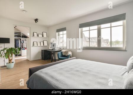 Das Bett mit grauer Decke befindet sich neben dem Schreibtisch mit Computer in einem geräumigen, sonnendurchfluteten Schlafzimmer mit Fenstern und Schranktür Stockfoto