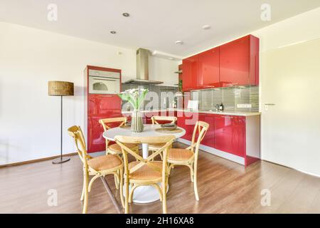 Moderne Innenausstattung der Küche mit roten Schränken und weißem Esstisch mit Blumen in Vase in zeitgemäßen Wohnung dekoriert Stockfoto