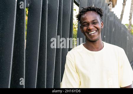Fröhlicher afroamerikanischer Mann mit lässigem hellgelben T-Shirt und kurzen Haaren, die am Zaun stehen und lachen, während sie die Kamera betrachten Stockfoto