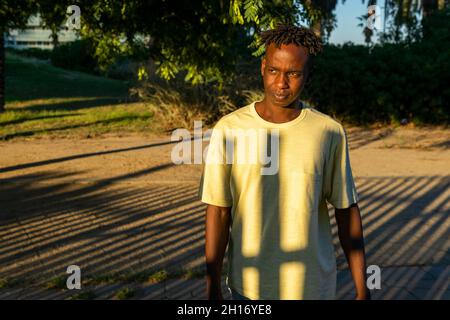 Junger afroamerikanischer Mann in legerer Kleidung, der auf einer Gasse im grünen Park steht und bei Sonnenuntergang nachdenklich davonschaut Stockfoto