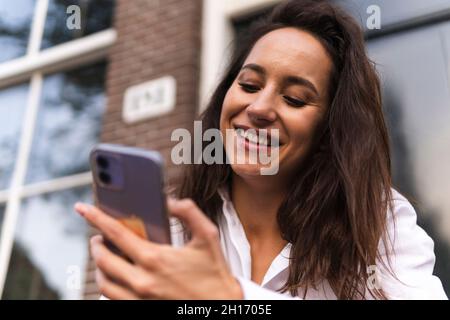 Nahaufnahme einer glücklichen jungen Frau in legerer Kleidung, die in der Nähe des Eingangs des Gebäudes steht und sich auf dem Handlauf stützt, während sie das Smartphone benutzt Stockfoto