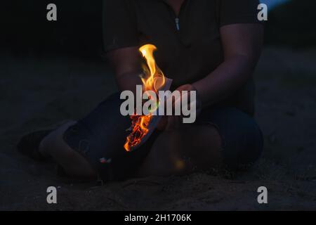 Eine Frau hält ein brennendes Stück Papier in ihrer Hand. Eine Erwachsene Frau sitzt nachts allein am Strand. Ein zerrissenes Blatt Papier aus einem Tagebuch mit Flammen. De Stockfoto