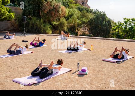 Eine Gruppe von Frauen, die nicht erkennbar auf Matten liegen und Bow Pose machen, während sie an sonnigen Tagen im Park mit grünen Bäumen Yoga mit einem Lehrer praktizieren Stockfoto