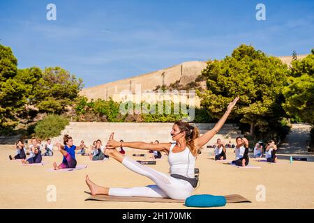 Seitenansicht einer flexiblen Lehrerin im aktiven Ohr, die während der Yoga-Gruppensitzung im Park sitzende, drehbare Hand bis zu großen Zehen aufführt und demonstriert Stockfoto