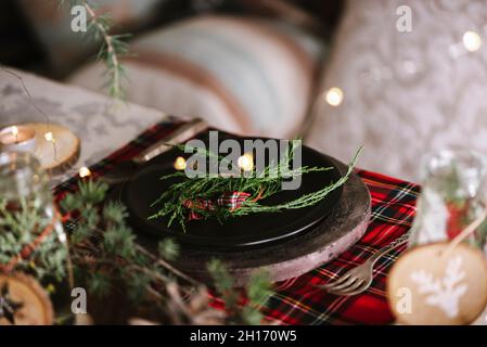 Weihnachtliche Tischeinstellung mit Kranz auf Keramikplatte auf rot karierter Tischdecke auf dem Hintergrund mit Lichtern Stockfoto