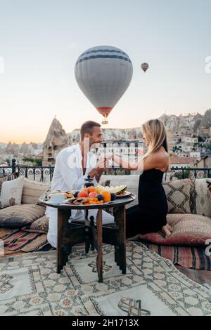 Mann küsst die Hand der Freundin, während er auf dem Dach in der Stadt mit Heißluftballons sitzt, die am Abendhimmel fliegen Stockfoto