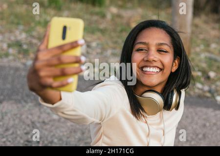 Glückliche ethnische Frau, die auf Longboard sitzt und Selbstporträt auf dem Handy macht Stockfoto