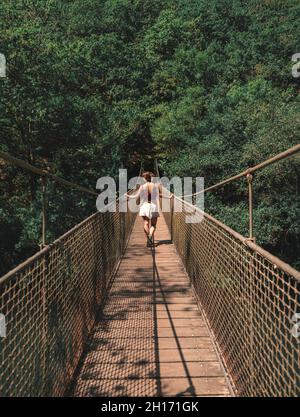 Rückansicht einer anonymen Forscherin, die an einem sonnigen Tag in Spanien auf einer Metallhängebrücke im Naturpark Fragas do Eume steht Stockfoto