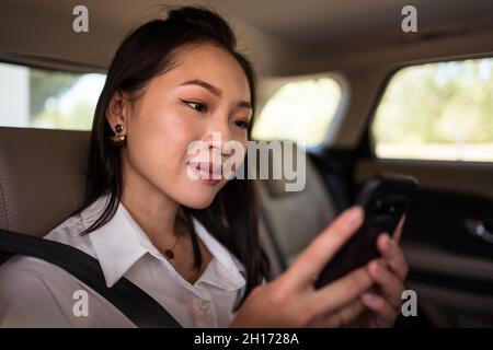 Seitenansicht einer asiatischen Beifahrerin in formeller Kleidung, die im Taxi auf dem Rücksitz durch das Mobiltelefon surft Stockfoto