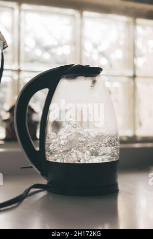 Transparenter Wasserkocher mit schwarzem Griff und Deckel und kochendem Wasser auf weißem Tisch in der Küche Stockfoto