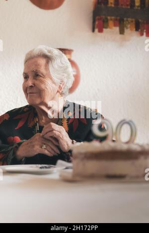 Gealterte Frau bläst Kerzen auf Geburtstagskuchen und klatscht dann die Hände, während sie den 90.. Jahrestag mit einem Verwandten zu Hause feiert Stockfoto